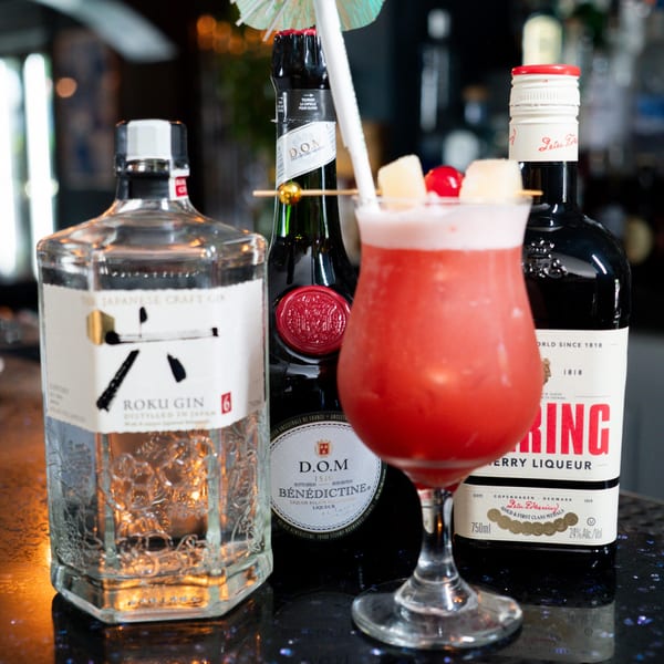 A vibrant red cocktail with pineapple and cherry garnishes, flanked by three liquor bottles on a bar.