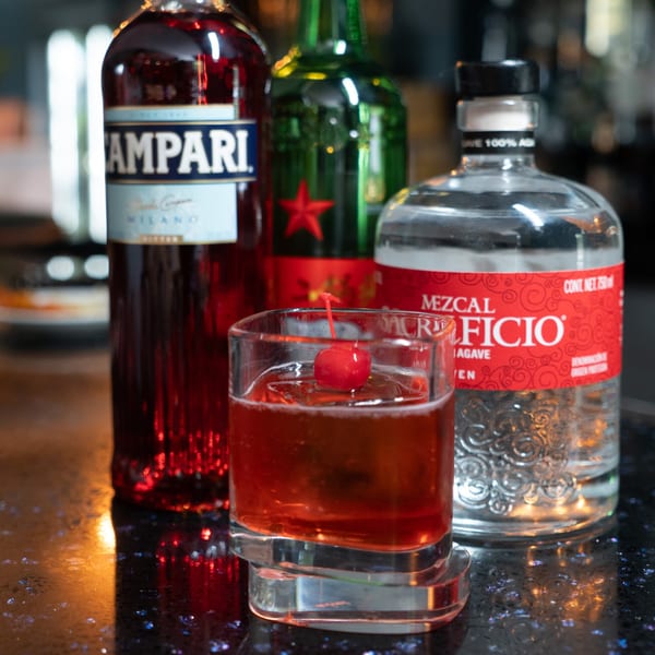 A red cocktail with a cherry garnish, surrounded by bottles of Campari, Heineken, and Mezcal.