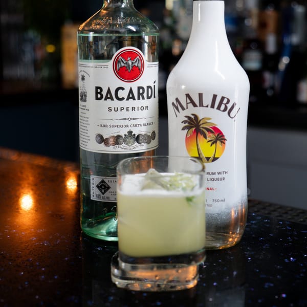Bacardi and Malibu rum bottles flank a cocktail with lime and ice on a dark bar.