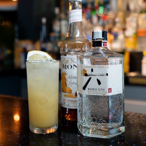 A tall glass of Roku Gin cocktail with a lemon slice, next to bottles of gin and honey syrup.