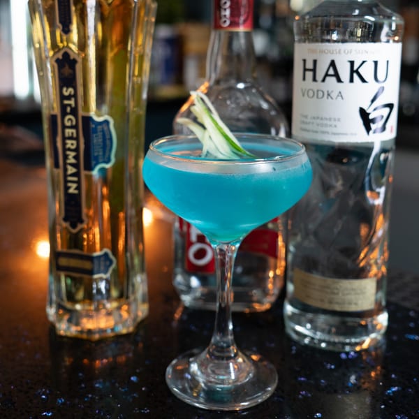 Blue cocktail garnished with cucumber slices, with bottles of St-Germain and Haku Vodka in the background.