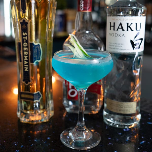 A vibrant blue cocktail garnished with cucumber slices, flanked by liquor bottles on a dark, sparkling surface.