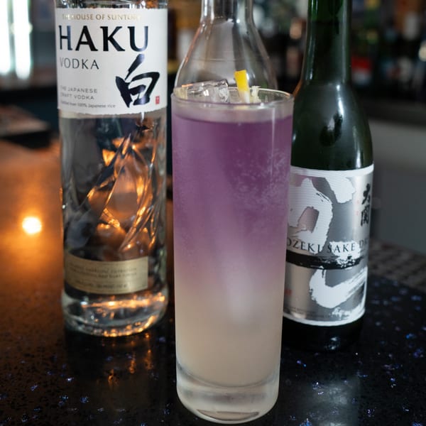 A purple and white layered cocktail with ice and lemon, flanked by Haku Vodka and Ozeki Sake bottles.