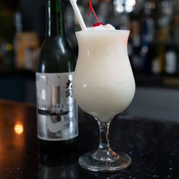 A frozen white cocktail with a cherry and straw next to a bottle of sake on a dark bar.