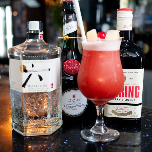 A red cocktail with pineapple and cherry garnishes, flanked by bottles of Roku Gin and liqueurs.