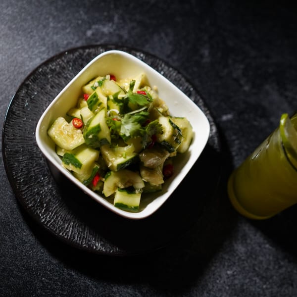 A white bowl of Asian-fusion cucumber salad with red chilies and cilantro, next to a green drink.