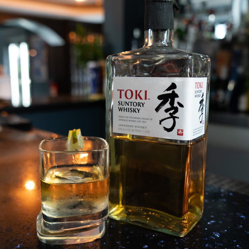 Toki Suntory Japanese Whisky bottle and a glass with ice and a lemon wedge on a bar.
