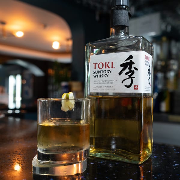 A bottle of Toki Suntory Whisky next to a glass with a drink and lemon peel on a bar.